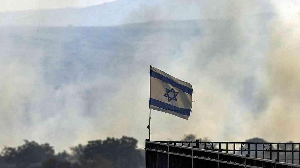 Israel alerta para aumento de ataques contra Hezbollah no Líbano | Uma bandeira israelense tremula em um poste enquanto nuvens de fumaça sobem de um incêndio em um campo depois que foguetes lançados do sul do Líbano caíram perto de Katzrin, nas Colinas de Golã anexadas por Israel, em 13 de junho de 2024, em meio a confrontos transfronteiriços entre as tropas israelenses e o Hezbollah