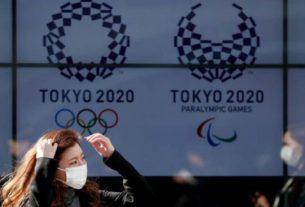 Mulher com máscara de proteção caminha em frente aos logos da Olimpíada e da Paralimpíada Tóquio